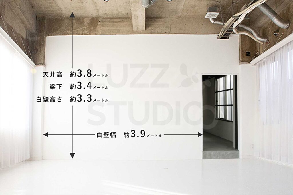 スタジオの白コンクリート床の部屋のサイズ詳細。天井高3.8メートル、梁下3.4メートル、白壁の有効幅3.9メートル。右側に隣接エリアへの入り口、両サイドに白いカーテンがある明るい内装