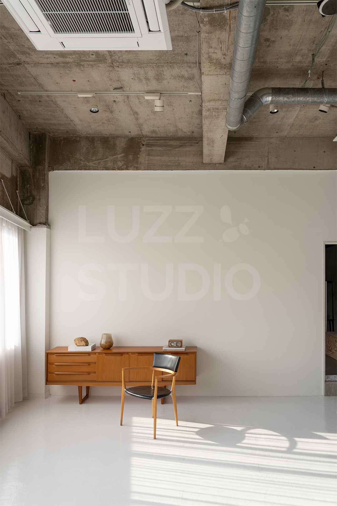 ラズスタジオの白コンクリート床の部屋にサイドボードと椅子が置かれた自然光差し込む室内写真